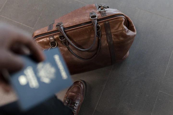 We look down at a passport held in our hand and below on the floor a brown leather travel bag.